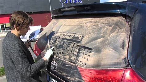 Menininkas Robertas Burdenas piešia portretą ant purvino automobilio naudojant specialų įrankį