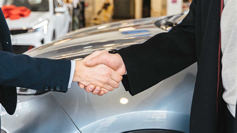 Teminė nuotrauka, vaizduojanti pirkėjo ir pardavėjo rankas, pasirašančias sutartį prie automobilio