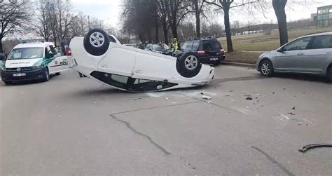 Tamsios spalvos automobilis apvirtęs ant stogo stovėjimo aikštelėje, aplink susirinkę žmonės