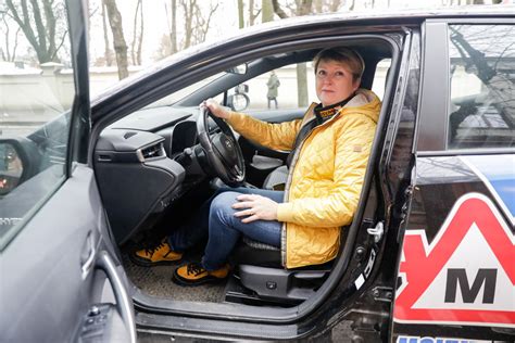 Teminė nuotrauka: suaugęs asmuo moko jauną žmogų vairuoti automobilyje, abu sėdi priekinėse sėdynėse, fone matyti kelias