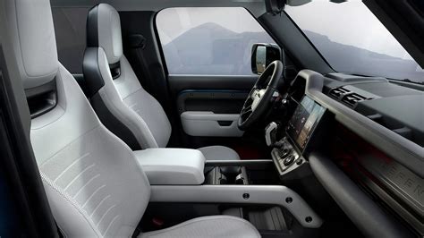 Land Rover Defender salono vaizdas, demonstruojantis centrinį ekraną, pavarų svirtį ir papildomą priekinę sėdynę.