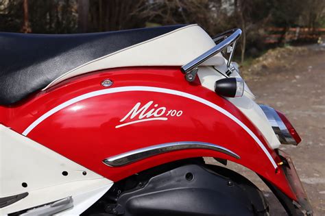 SYM Mio 100 motoroleris retro stiliaus, su apvaliu priekiniu žibintu ir chromo detalėmis.