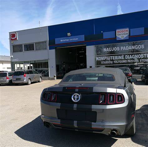 Teminė nuotrauka: žmogus, rankoje laikantis automobilio detalę, fone - įrankiai ir automobilių servisas